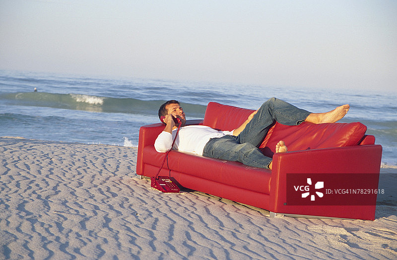 男人躺在沙滩边的沙发上玩手机图片素材