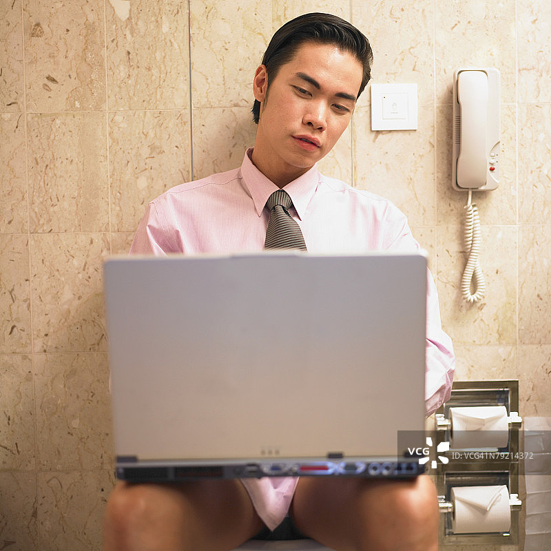 年轻商人坐在马桶上用笔记本电脑工作。图片素材