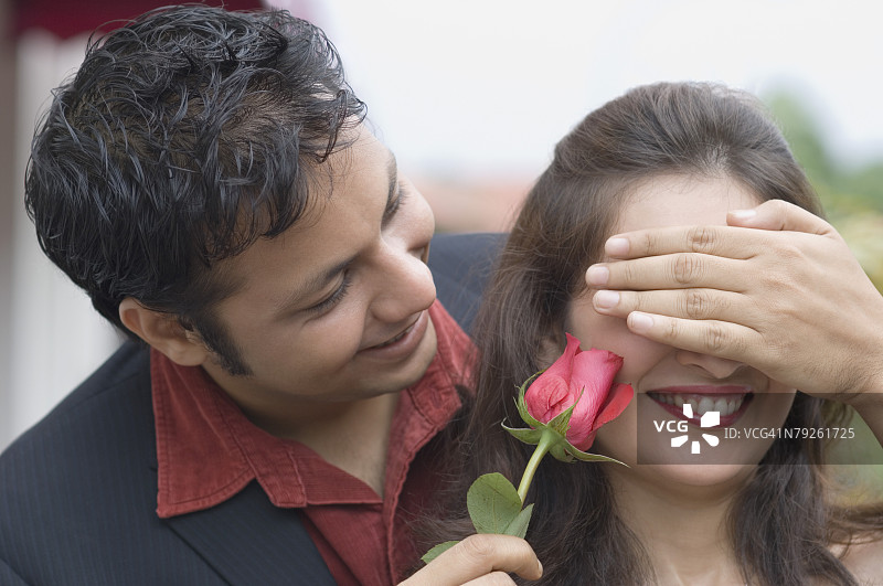 一位年轻男子用手遮住一位年轻女子的眼睛，并手持玫瑰花的特写镜头图片素材