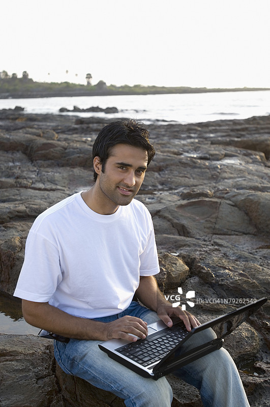 年轻男子微笑着将笔记本电脑放在腿上，位于印度马哈拉施特拉邦孟买的马德岛。图片素材