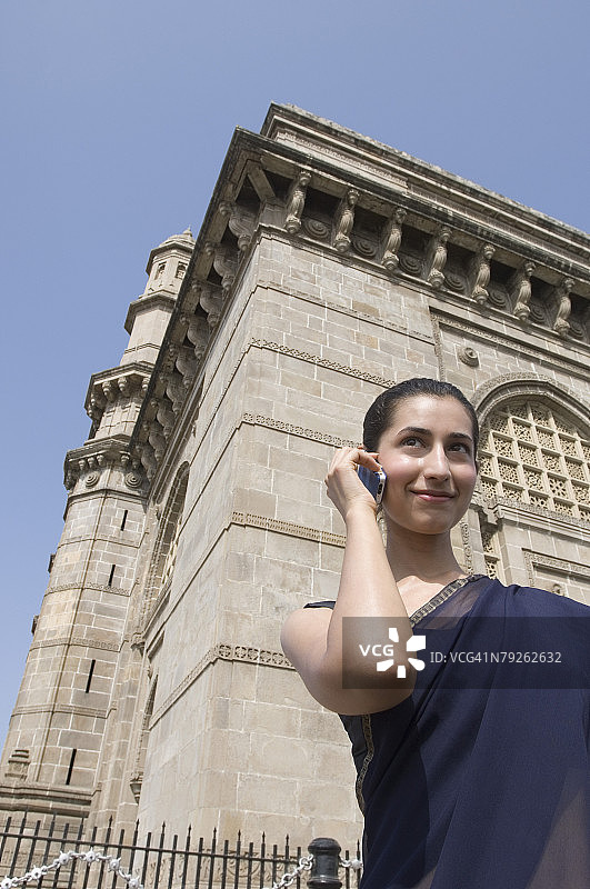 印度孟买印度门，年轻女子站在纪念碑前用手机交谈的低角度视图图片素材
