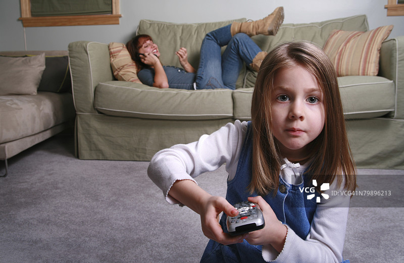 一个五岁女孩在换电视频道，而她的保姆/年轻母亲在背景中忽略了她图片素材