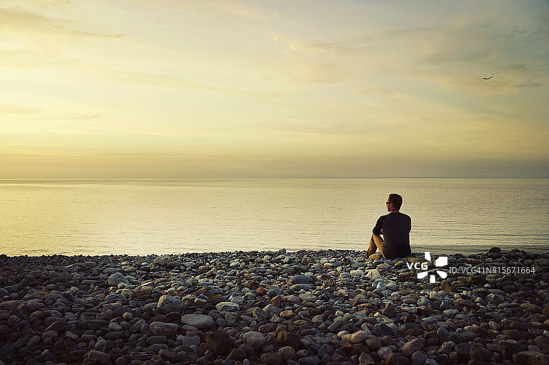 男人独自坐在鹅卵石海滩上，眺望着平静的海面图片素材