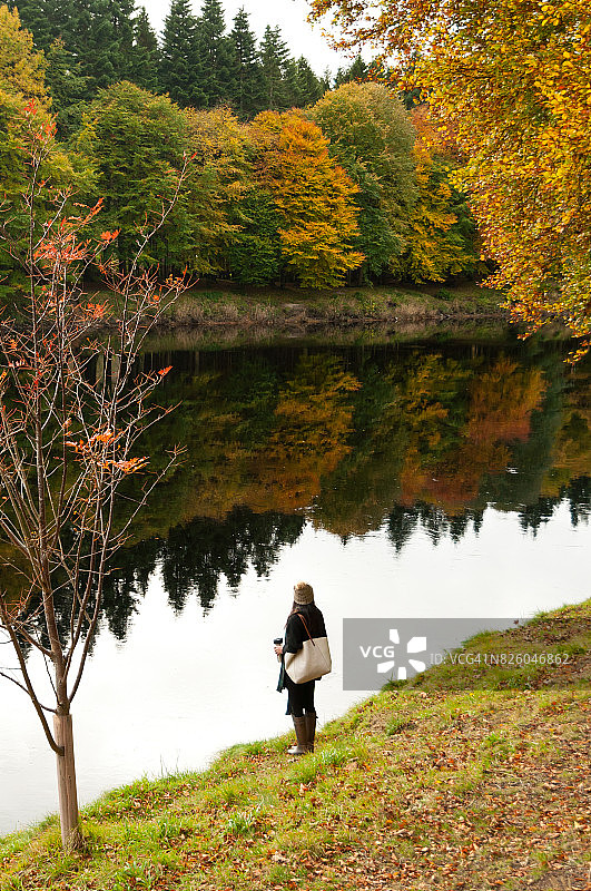 戴着羊毛帽站在平静的河流旁，河面有彩色倒影的亚洲女孩图片素材