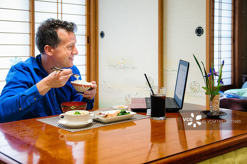 在日式房间里吃早餐并对着笔记本电脑微笑的白人男子图片素材