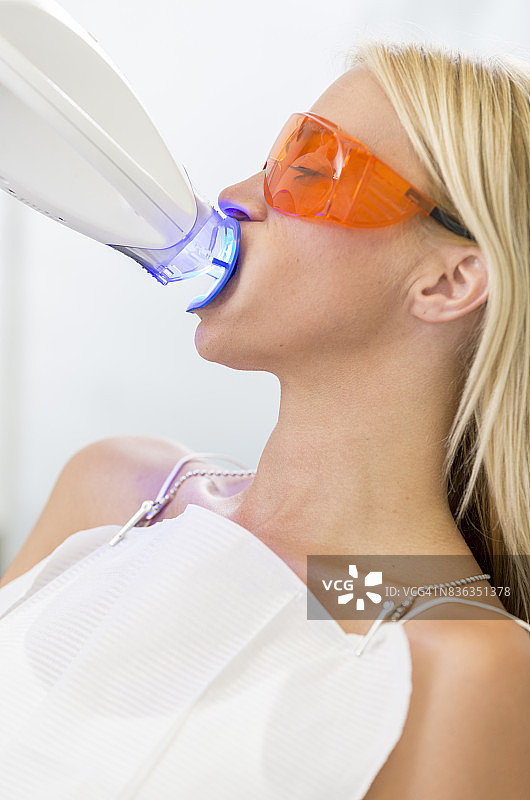 美容牙科护理 - 金发碧眼的女性躺在牙医诊室的椅子上，张开嘴进行牙齿美白，同时戴着防护眼镜和唇部牵开器，插入美白装置发出紫外线。图片素材