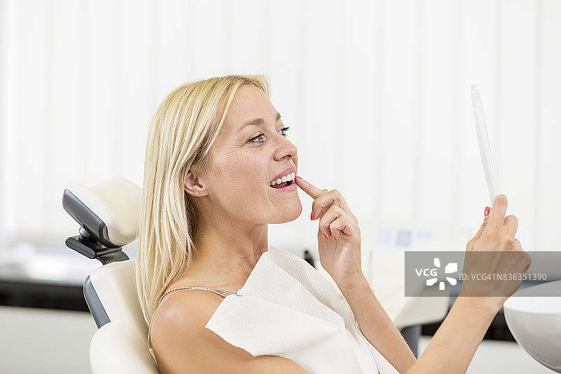 坐在椅子上的金发女性患者正在用手镜检查牙齿清洁和美白后的治疗效果图片素材