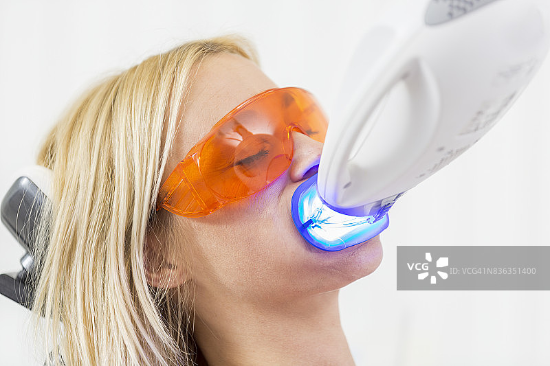 美容牙科护理 - 金发碧眼的女性躺在牙医诊室的椅子上，张开嘴进行牙齿美白，同时戴着防护眼镜和唇部牵开器，插入美白装置发出紫外线。图片素材