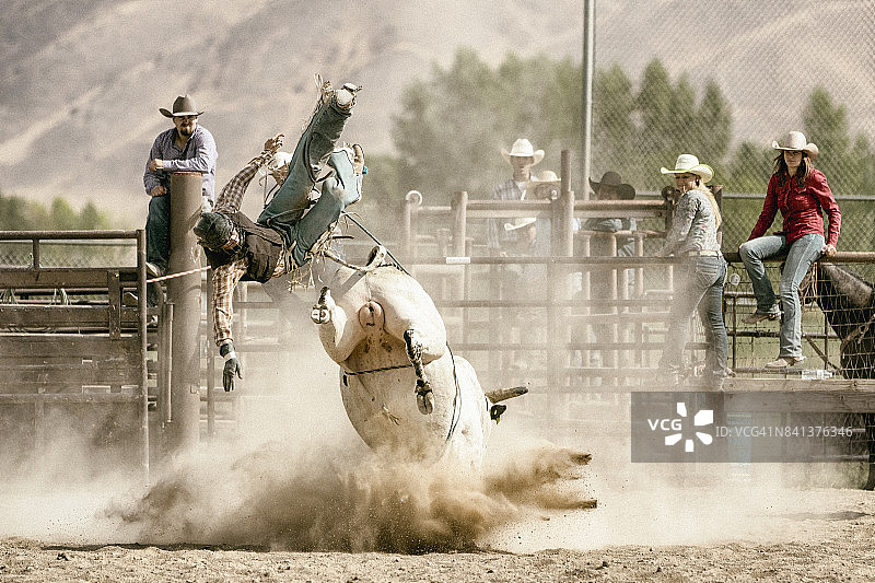 一个年轻的牛仔赤膊骑在一头烈性公牛上，一群牛仔在背景中观看图片素材