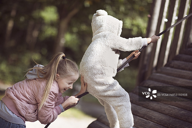 欢笑的女孩推着穿着熊套装的孩子爬上操场绳索图片素材