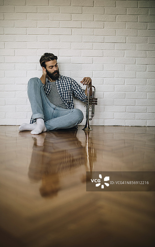 坐在镶木地板上吹小号的年轻络腮胡男士图片素材