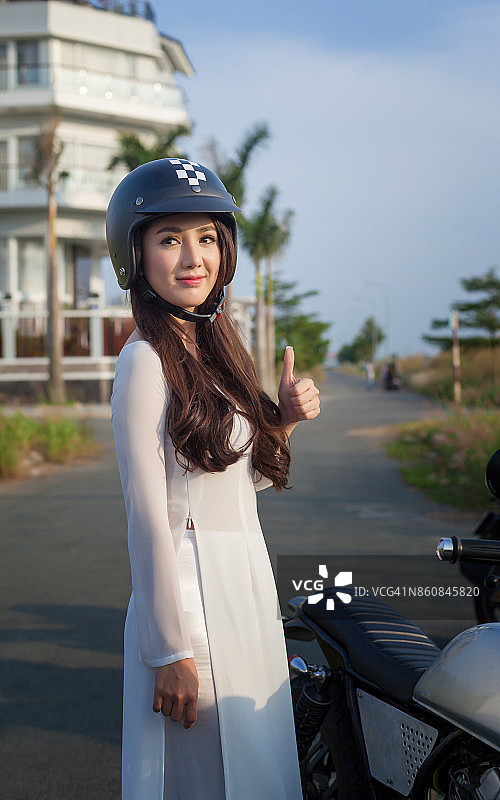 年轻女孩穿着奥黛，戴着头盔，站在摩托车旁，伸出一根手指图片素材