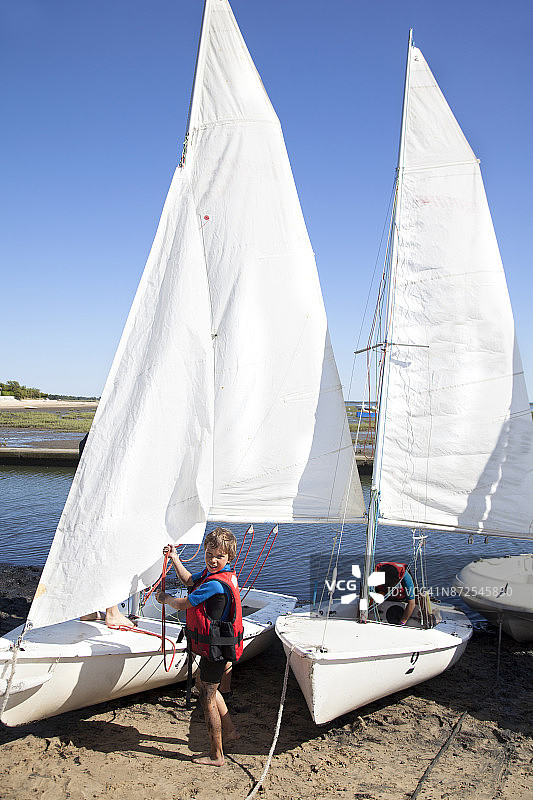 穿着潜水服的男孩站在小艇旁，将绳子穿过帆图片素材
