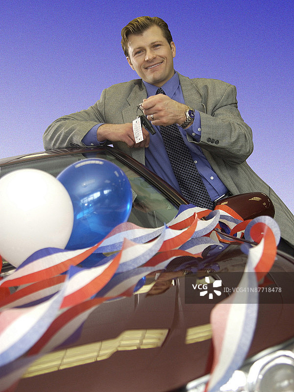 一位靠在汽车上，拿着钥匙看着观众的年轻白人男汽车销售员图片素材