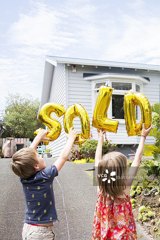 站在房子前，拿着金色“已售”气球的男孩和女孩图片素材