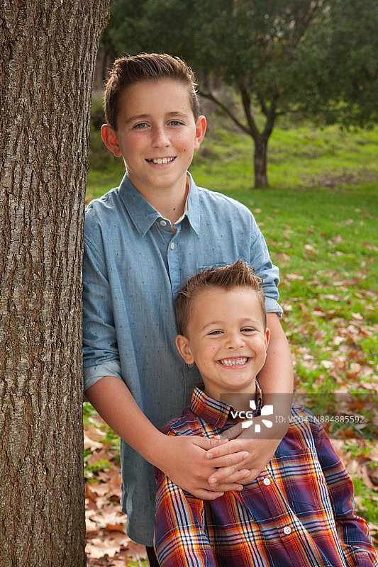 哥哥和弟弟在户外自然公园里微笑，靠在树上看着镜头图片素材
