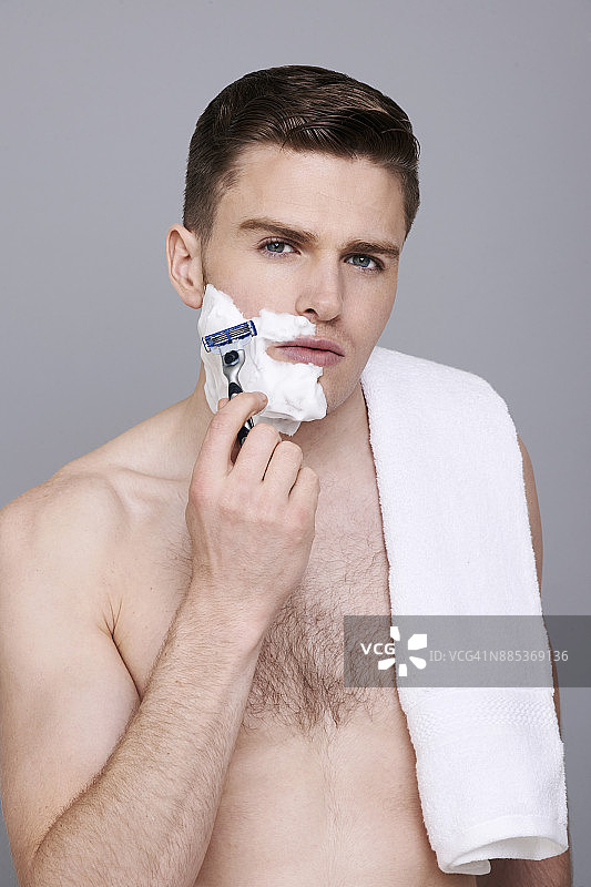 男人肩搭毛巾，半张脸涂剃须膏，手持剃须刀，表情严肃图片素材