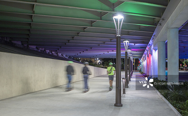 夜晚，三个人骑着滑板车在灯火通明的便道上通勤图片素材