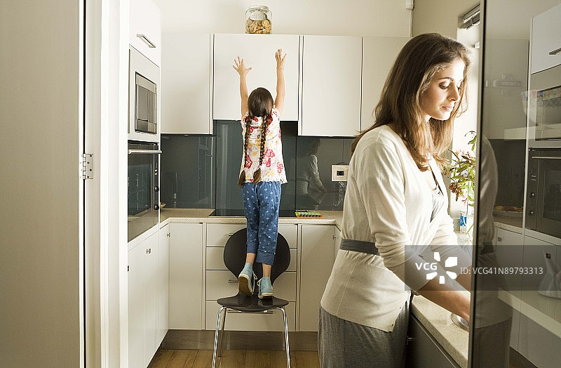 在厨房里，女儿伸手去够饼干罐，母亲在一旁图片素材