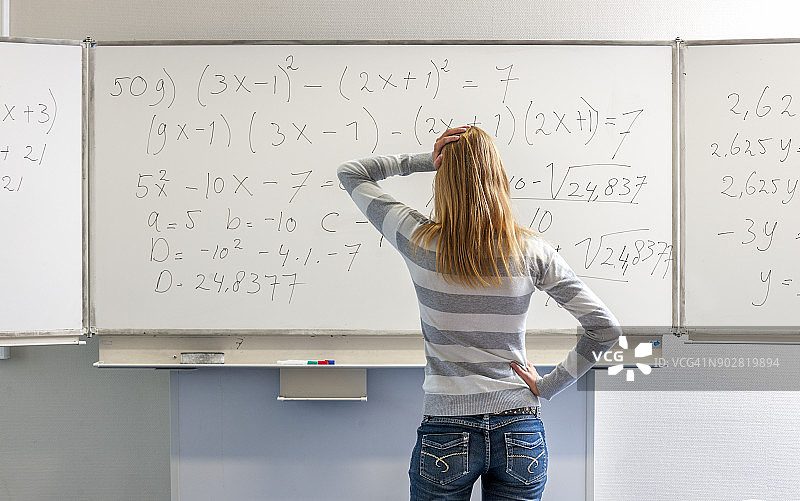 一个困惑沉思的金发高中女生站在白板前，手托下巴，看着白板上的数学公式图片素材
