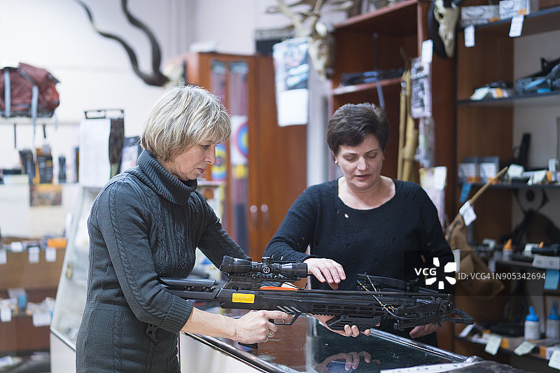 一位有魅力的金发中年女人在狩猎商店里，在女销售员的帮助下，检查十字弓图片素材