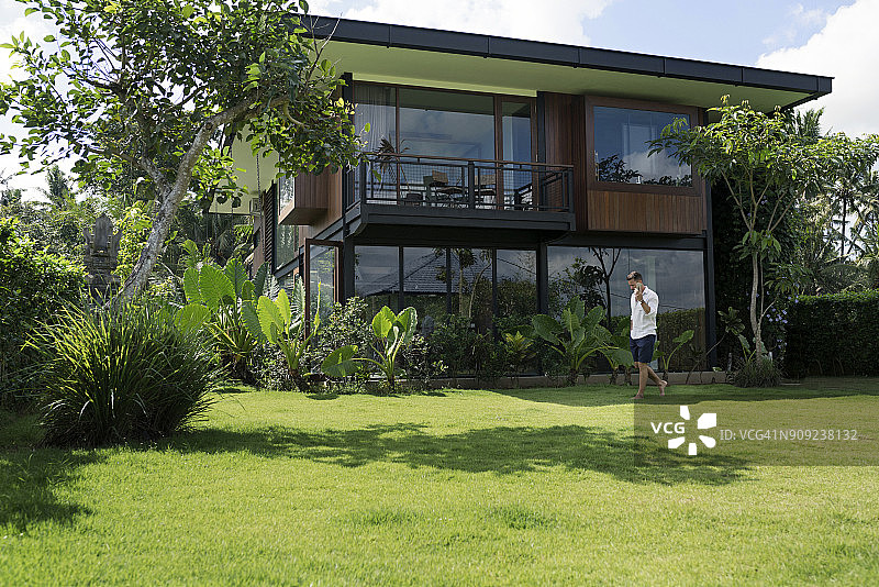中年男人在现代别墅前的花园里散步，使用智能手机图片素材