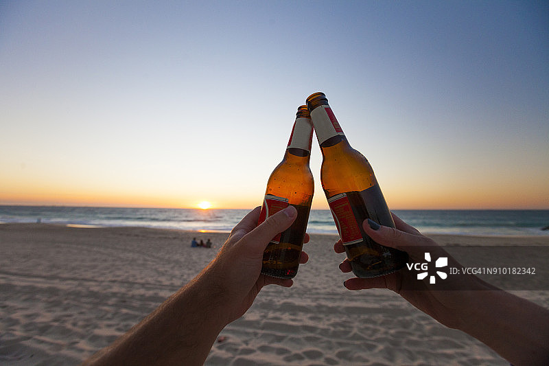 两人在澳大利亚西澳大利亚珀斯海滩日落时分用啤酒瓶进行庆祝祝酒图片素材