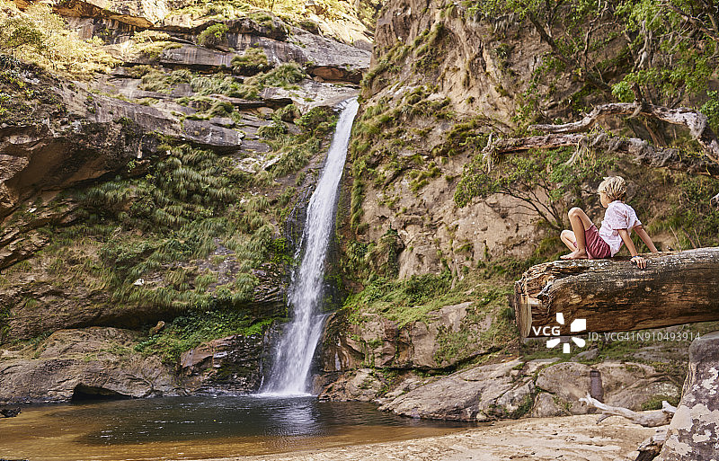 男孩坐在原木上看着瀑布，玻利维亚圣克鲁斯萨迈帕塔图片素材
