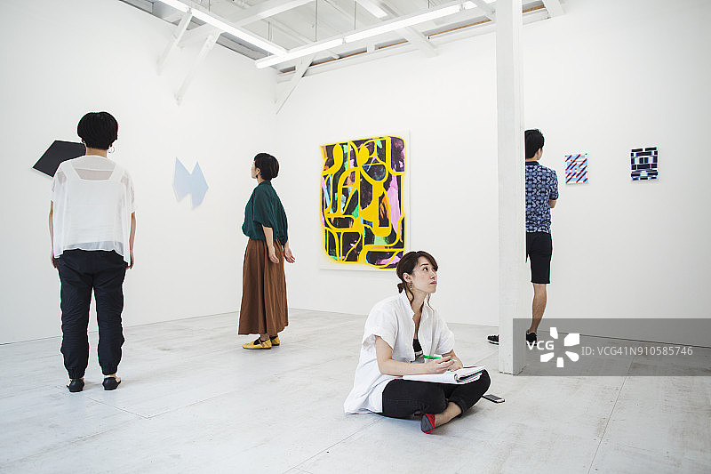 在美术馆地板上，用笔和纸看着现代画作的黑发女人图片素材