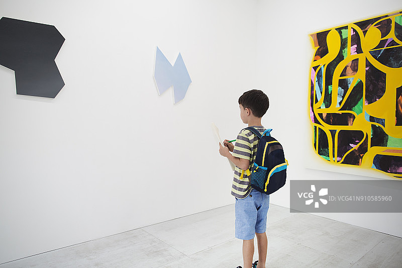 在美术馆里，留着黑色短发，背着背包，拿着纸笔，看着现代绘画的男孩图片素材