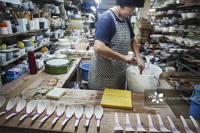 女人在日式瓷器作坊工作，将白色瓷勺浸入釉料桶中图片素材