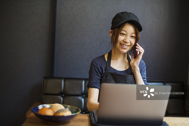 在面包店工作的戴棒球帽的女人，坐在桌前使用笔记本电脑和手机图片素材