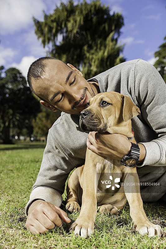 年轻男人躺在草地上，在公园里拥抱他的小狗图片素材