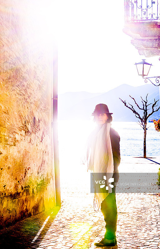 戴着帽子和围巾站在阳光明媚的古老街道上的优雅女人图片素材