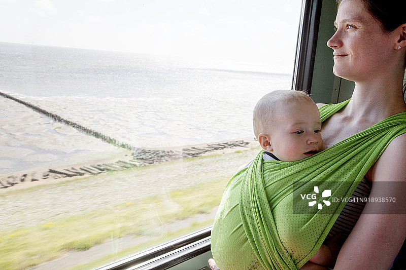 母亲用背巾抱着稍大的宝宝望向窗外图片素材
