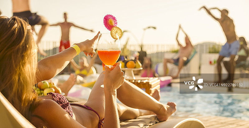 女子手持鸡尾酒躺在泳池边，日落时分人们在泳池中及周围跳舞图片素材