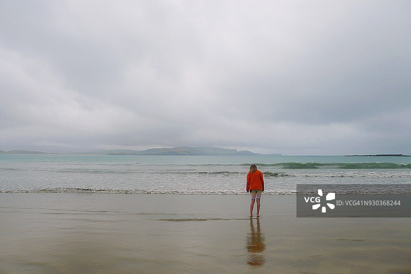 雨天独自在海滩的女孩，远处水中有赫氏海豚，地点：库里奥湾的波波斯湾，卡特林斯图片素材