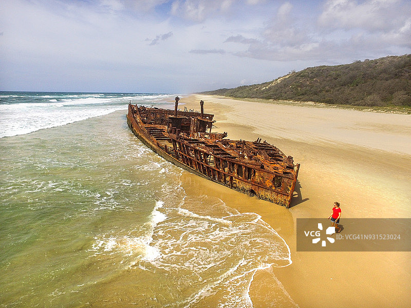 海洋边的沉船的航拍无人机视角，一个人穿着红色衣服走在长长的海滩上。图片素材