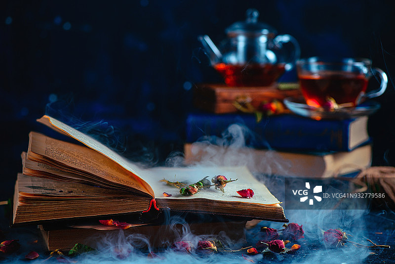 打开的书本，神秘的烟雾和玫瑰花瓣，茶杯在黑暗背景中失焦，魔法书和现代魔法概念图片素材