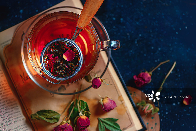 打开的书上的茶漏的特写。玫瑰花瓣草药茶。黑色饮品摄影概念。图片素材