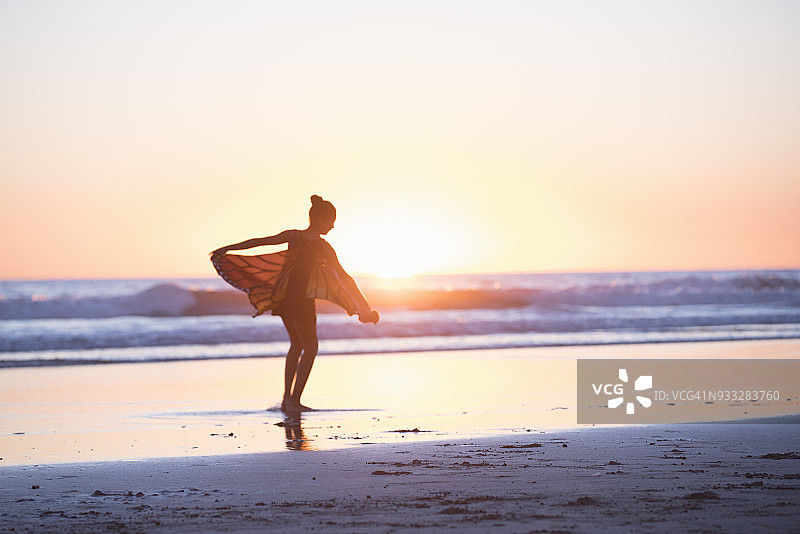戴着蝴蝶翅膀在海滩日落时跳舞的小女孩图片素材