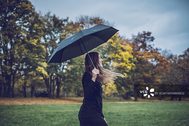 快乐的年轻女人打着雨伞在秋日公园散步图片素材