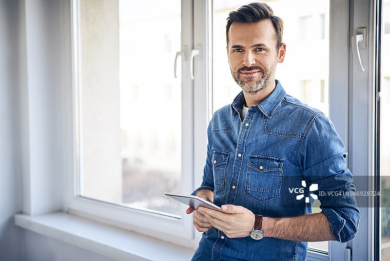 笑容满面的男人在窗边拿着平板电脑图片素材