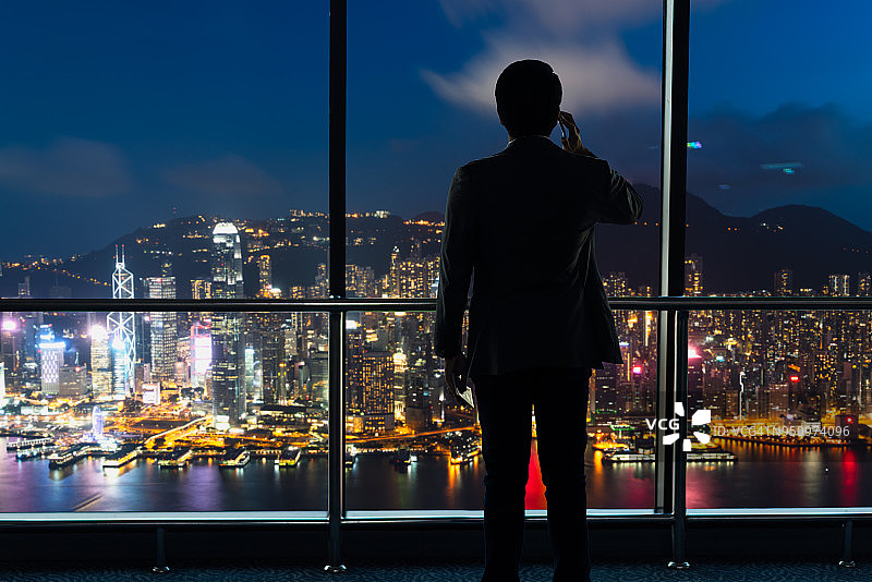 夜晚，年轻商人独自站在办公室的窗前使用手机图片素材