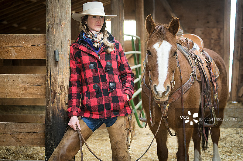 穿着红色格子夹克、牛仔帽的女牛仔在谷仓里牵着马缰绳图片素材