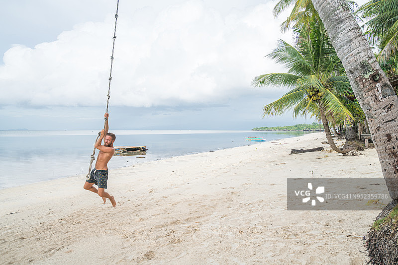 年轻男子在海滩上付款，棕榈树上有秋千绳图片素材
