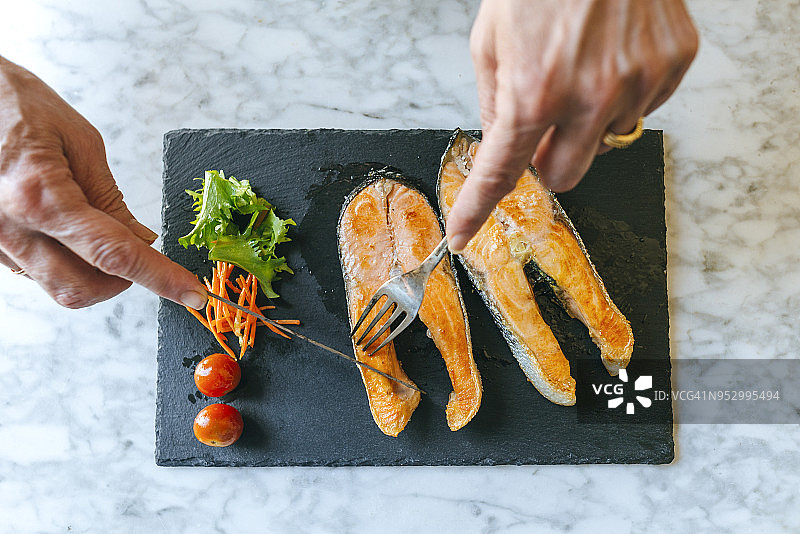 女子在盘中切割烤三文鱼片的手部特写，配有沙拉图片素材