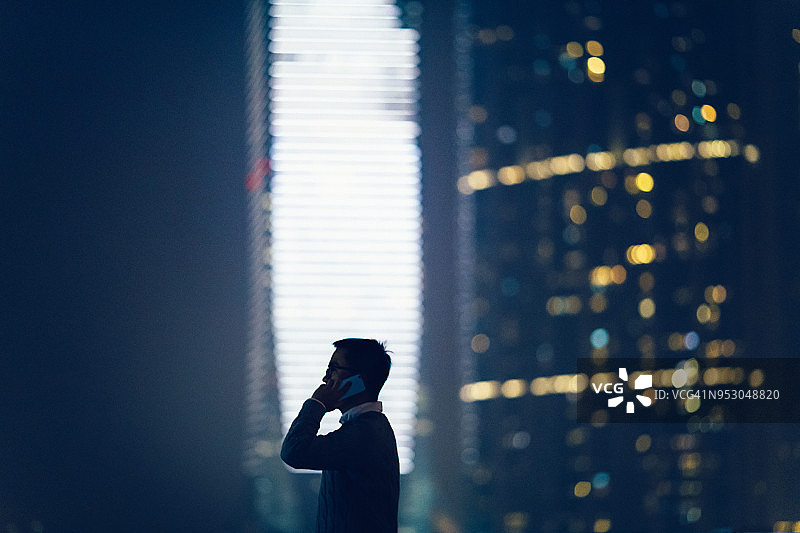 忙碌的商人在夜晚对着灯火通明的现代金融摩天大楼与客户通电话图片素材