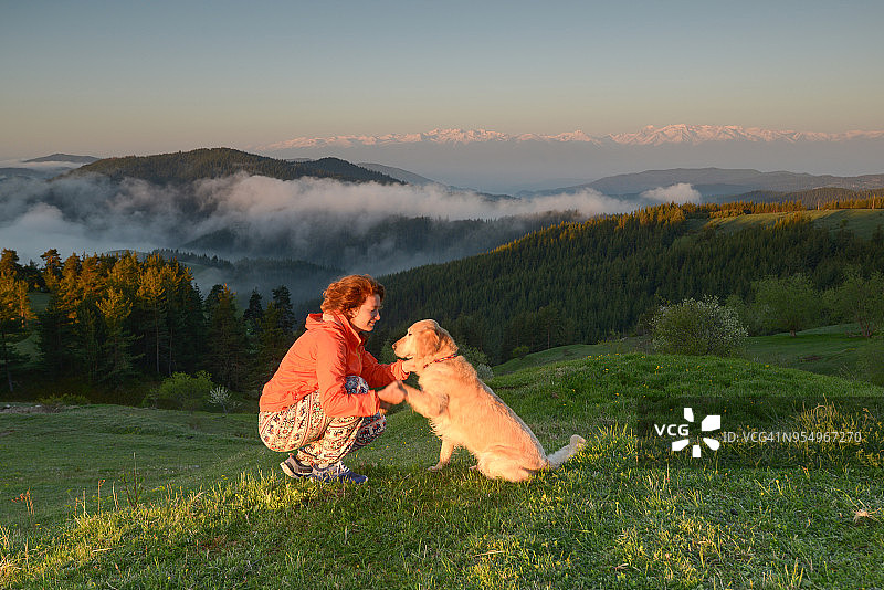 年轻女人和金毛猎犬在山间草地日出时图片素材