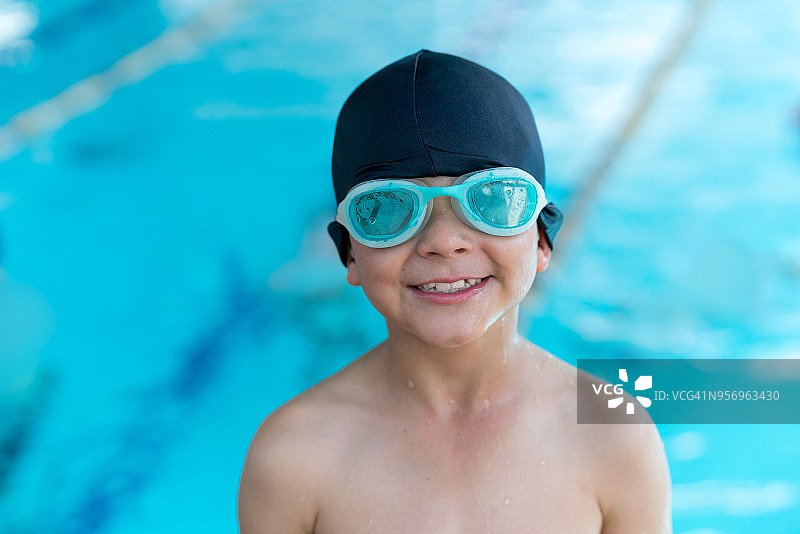 快乐的可爱男孩戴着泳镜和泳帽在游泳池里看着镜头，非常高兴图片素材